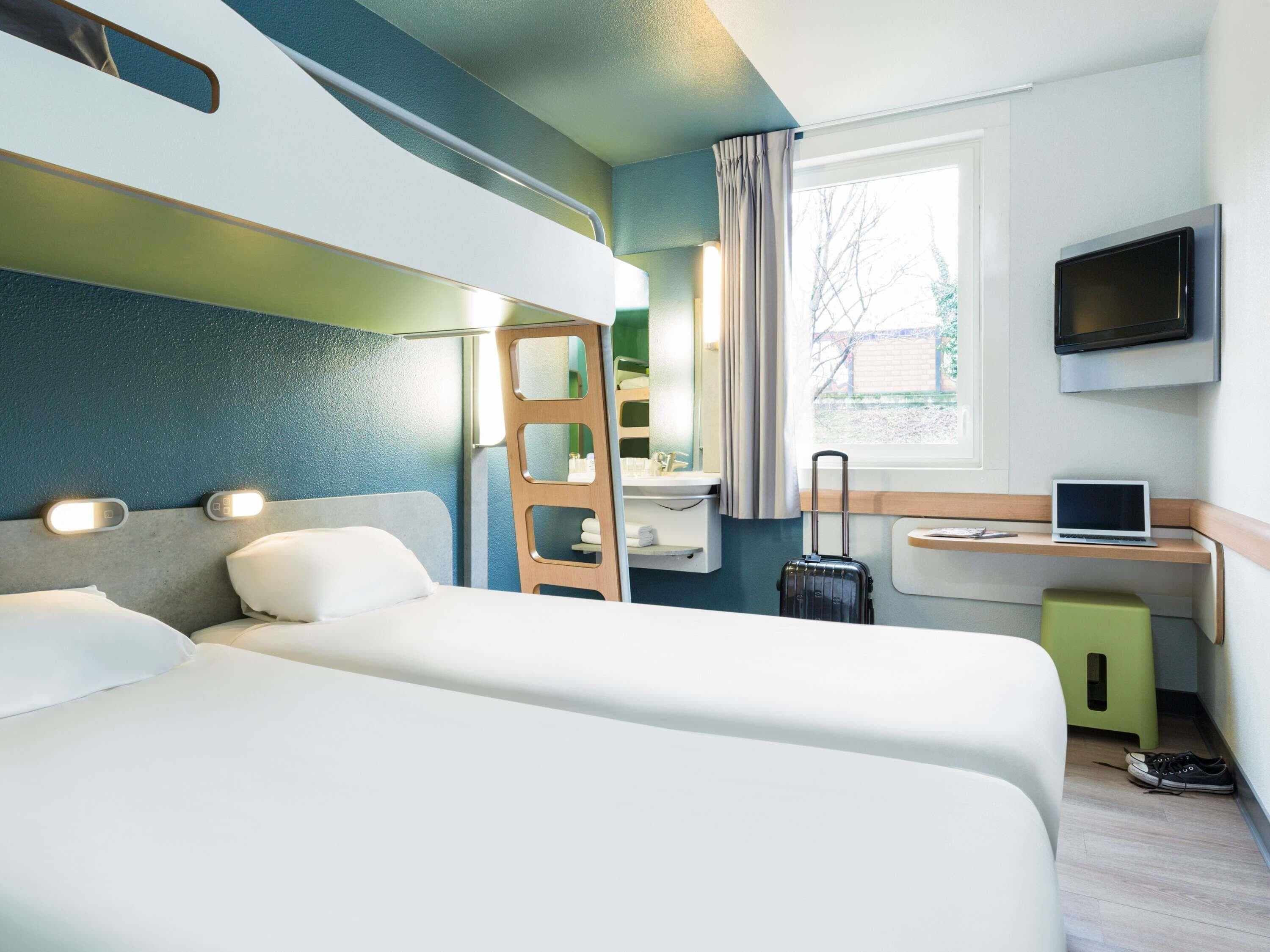 Quarto de hotel com duas camas, uma mesa com um laptop, uma televisão e uma mala.