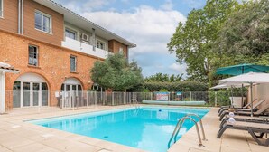 A rectangular swimming pool with a metal ladder, surrounded by a tiled deck. There are lounge chairs and umbrellas, and a brick building with windows and a balcony in the background. - Park & Suites Toulouse Colomiers (Toulouse)