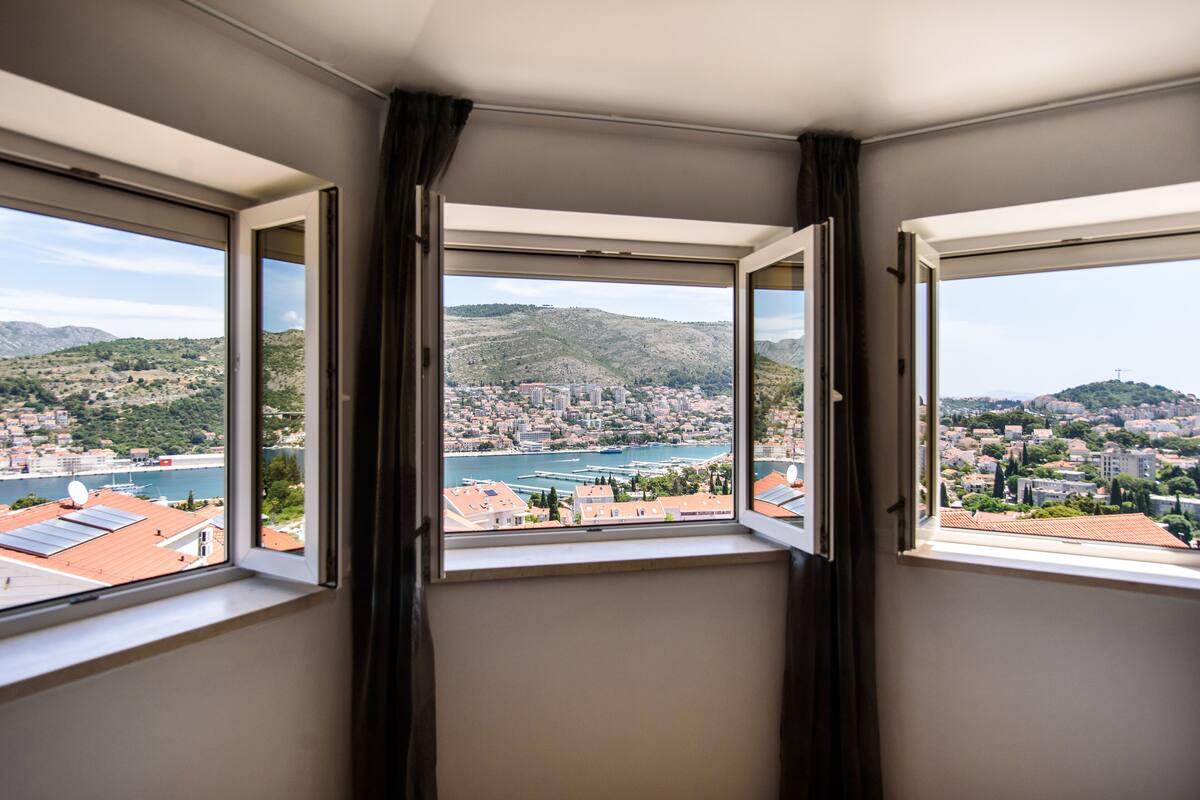 Quarto com janela panorâmica para uma paisagem costeira com colinas e um porto.