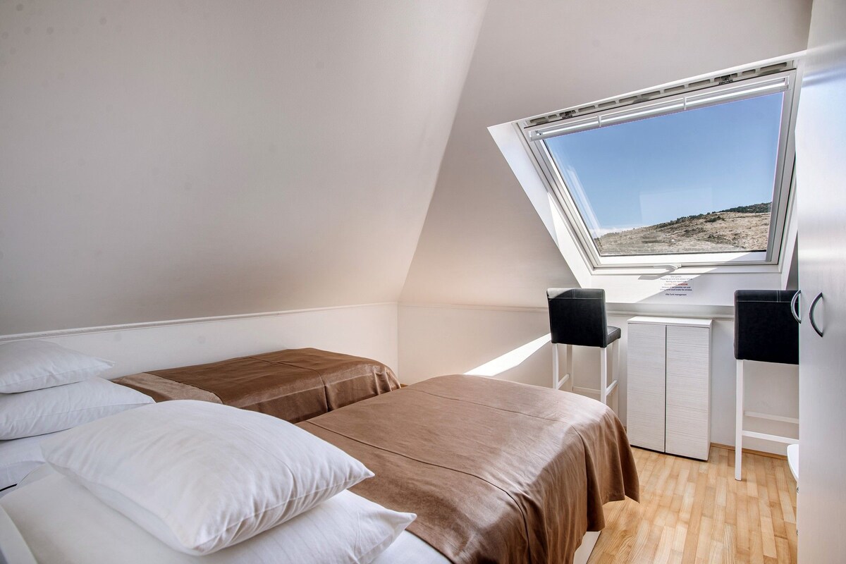 Quarto com teto inclinado, um claraboia, duas camas com diferentes roupas de cama, uma escrivaninha com cadeira e um pequeno armário.