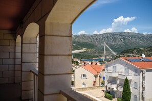 A balcony with stone arches overlooking a coastal town, a cable-stayed bridge, and a mountainous backdrop.