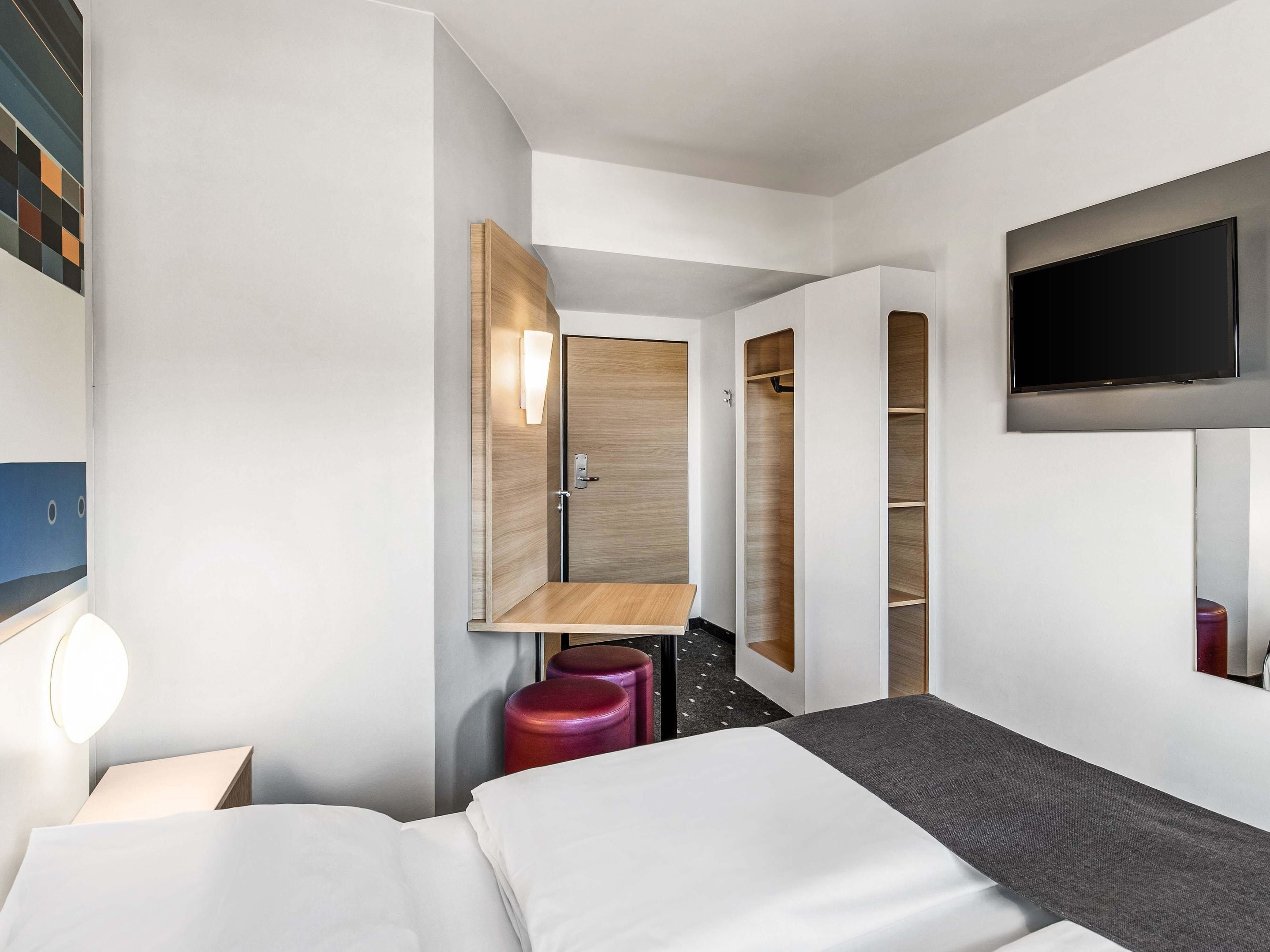 Ein modernes Hotelzimmer mit Bett, Wandfernseher, Holzschrank und kleinem Tisch mit Stuhl.
