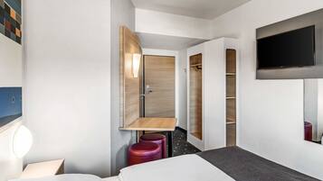 Ein modernes Hotelzimmer mit Bett, Wandfernseher, Holzschrank und kleinem Tisch mit Stuhl.