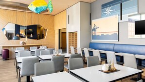 Un espace de restauration moderne, doté de plusieurs tables et chaises, d’une grande décoration représentant des poissons et d’un mur orné d’une boussole et d’une œuvre d’art représentant un paysage urbain.