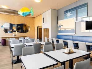 Een moderne eetruimte met meerdere tafels en stoelen, een grote visdecoratie en een muur met kompas en stadsgezicht kunstwerk.