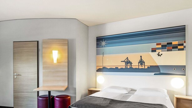 Habitación de hotel con cama, dos mesitas de noche, una lámpara de pared, un adorno en la pared y una puerta.