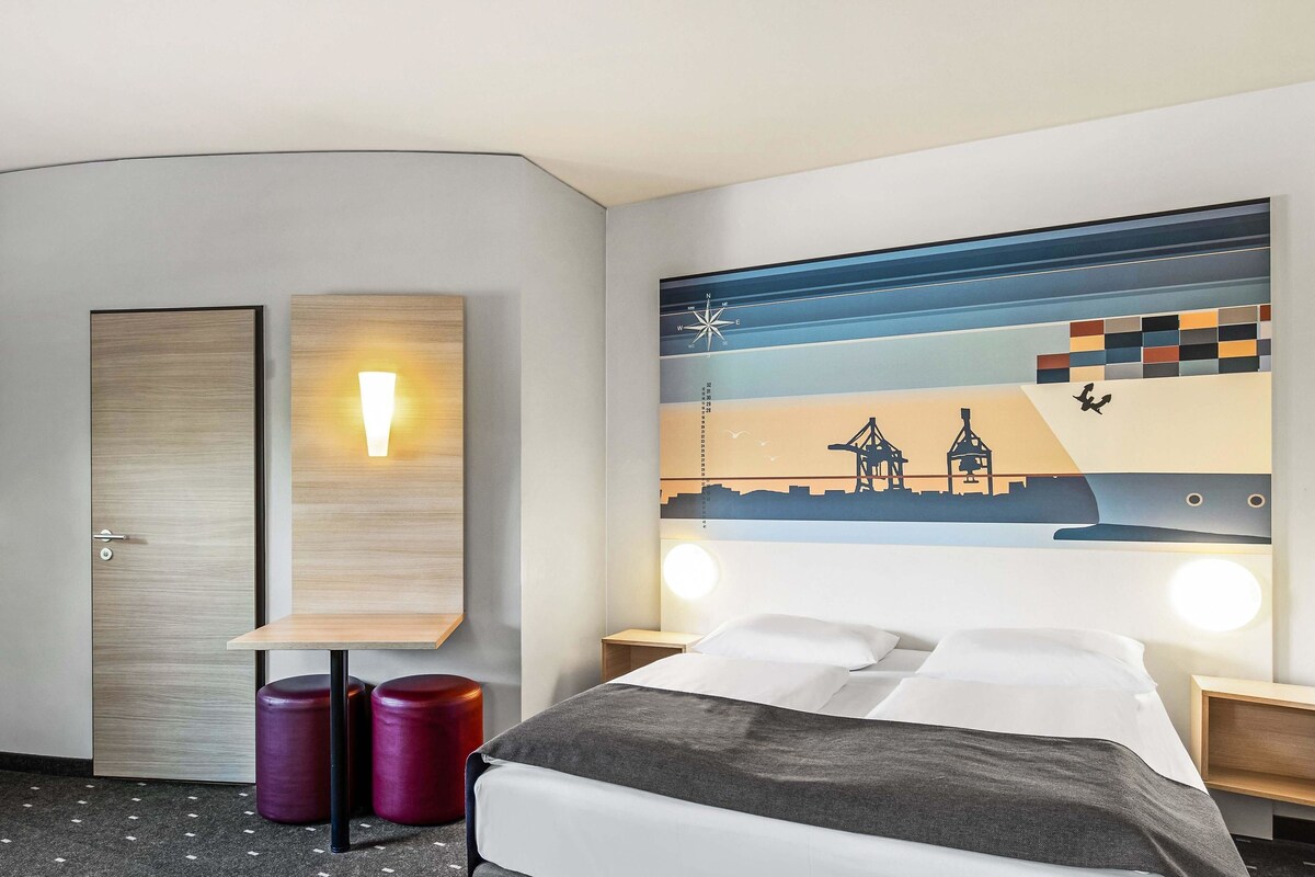 A hotel room with a bed, two bedside tables, a wall-mounted light, a decorative wall piece, and a door.