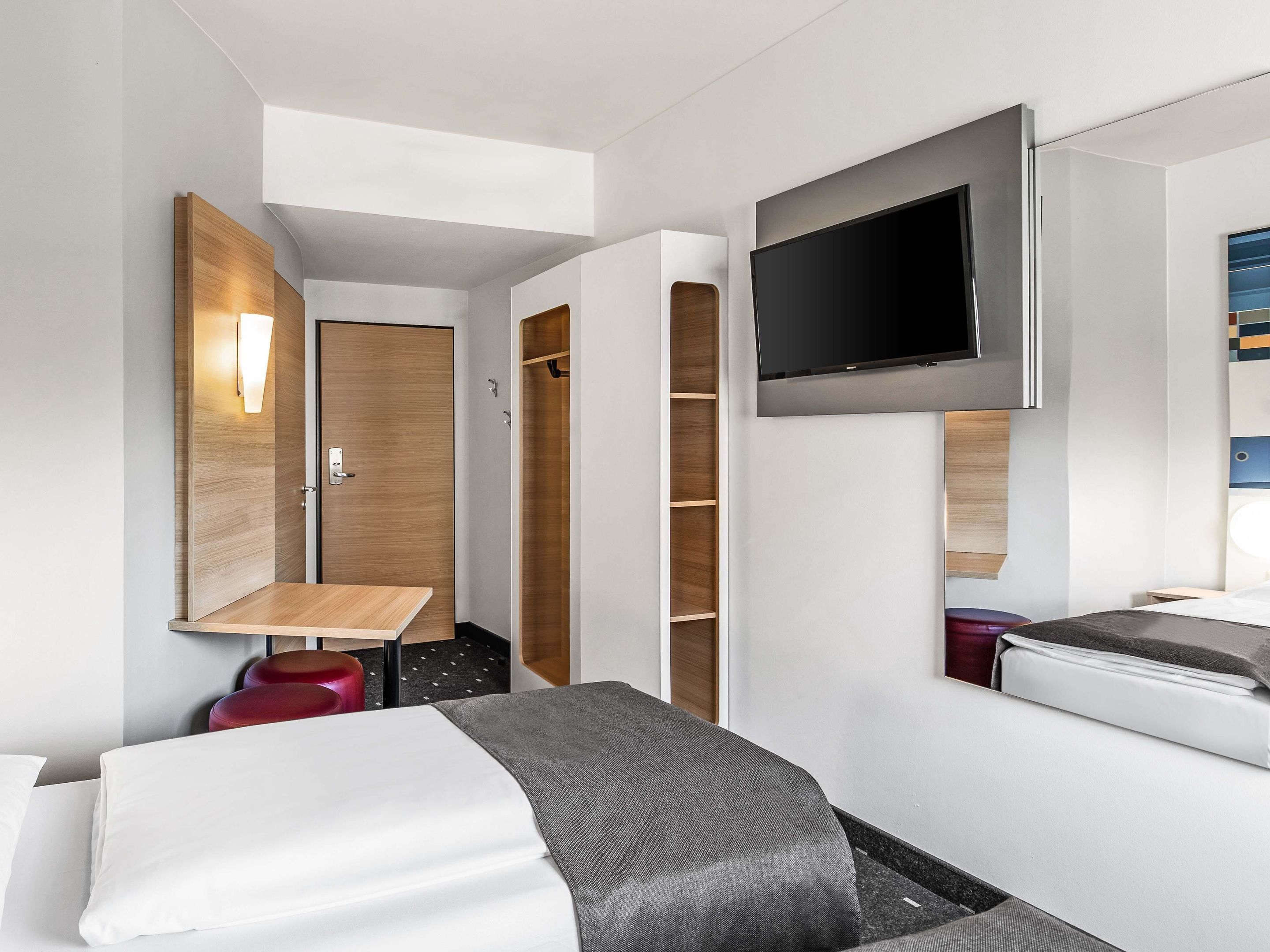A modern hotel room with a bed, a TV mounted on the wall, a wooden wardrobe, and a small table.