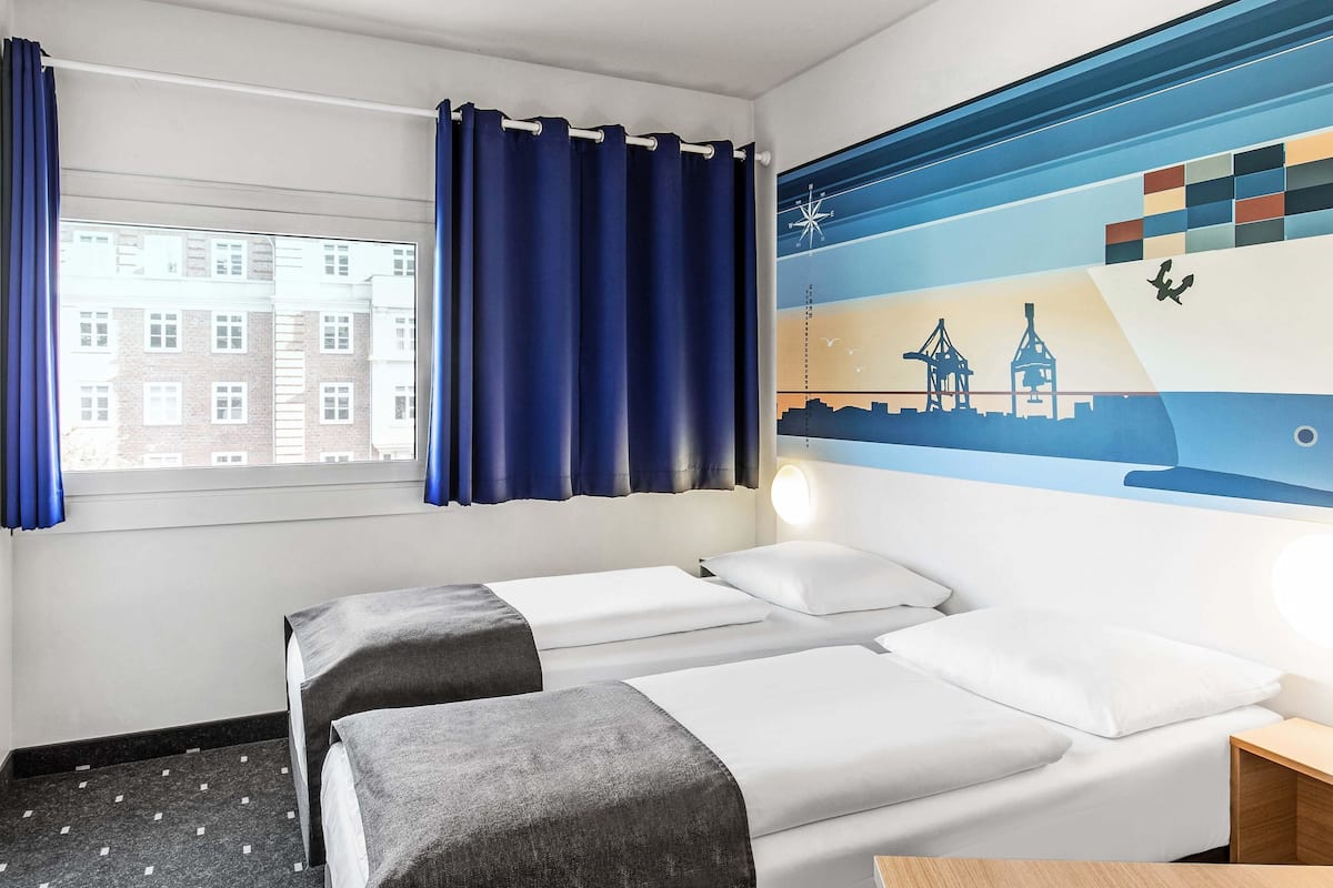 Une chambre d’hôtel avec deux lits, une fenêtre avec des rideaux, une fresque murale et une table de chevet.