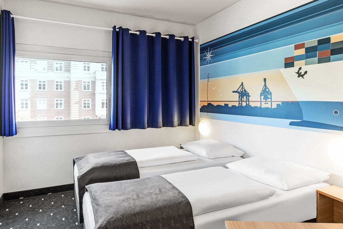 A hotel room with two beds, a window with curtains, a wall mural, and a nightstand.