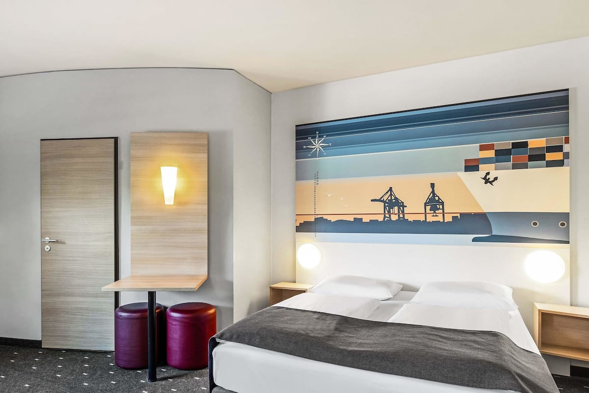Une chambre d’hôtel comprenant un lit, deux tables de chevet, une lampe fixée au mur, un élément décoratif mural et une porte.