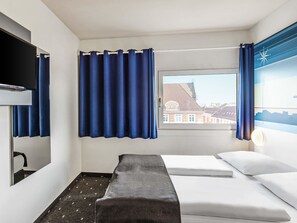 Een hotelkamer met een bed, een televisie, een raam met gordijnen en uitzicht op gebouwen.