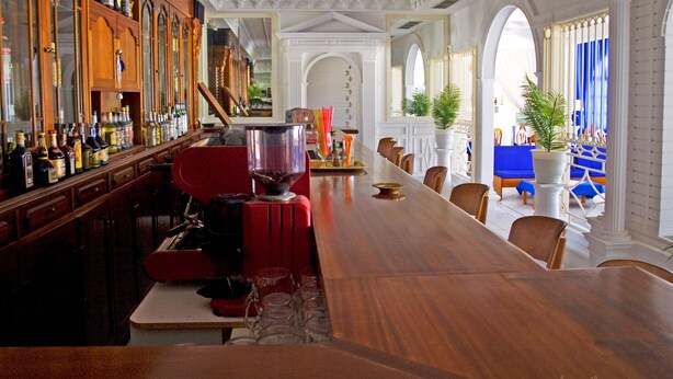 A bar area with a long wooden counter, shelves with bottles, and a coffee machine. - Parthenon (Ios)