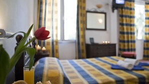 A bedroom with a striped bedspread, a bedside table with a lamp, a glass of orange juice, and a red flower. - Hotel Galaxias (Corfu)