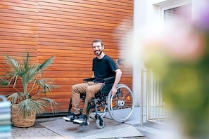 A person in a wheelchair outside a building with wooden paneling, a potted plant, and a metal gate.