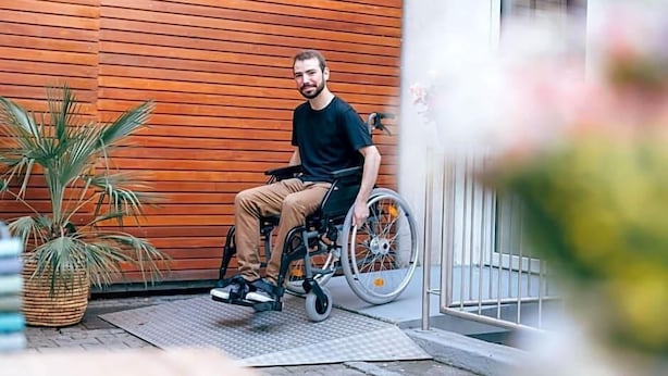 Una persona su sedia a rotelle di fronte a un edificio con rivestimento in legno e un cancello metallico.