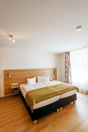 A hotel room with a wooden headboard, a bed with white linens, a bedside table, and a window with curtains.