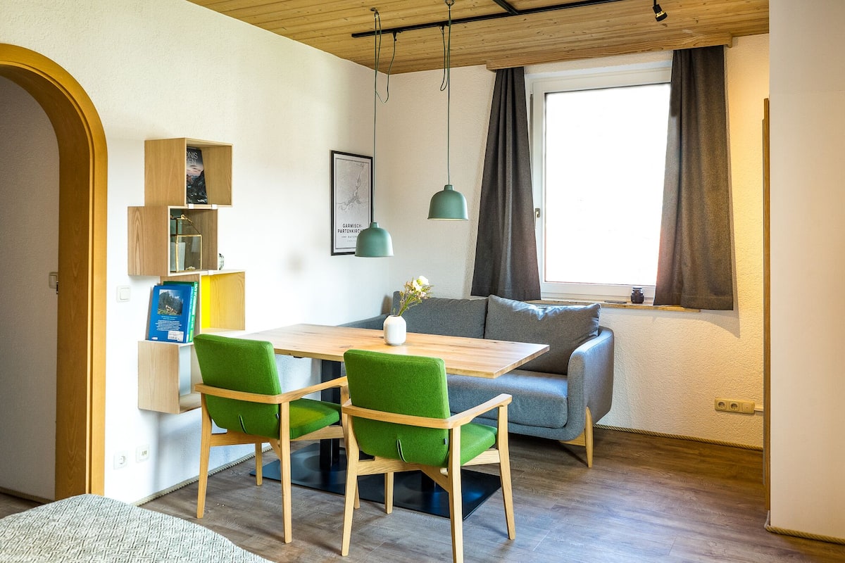 A cozy room with a wooden ceiling, a dining table with green chairs, a grey sofa, and a window with curtains.
