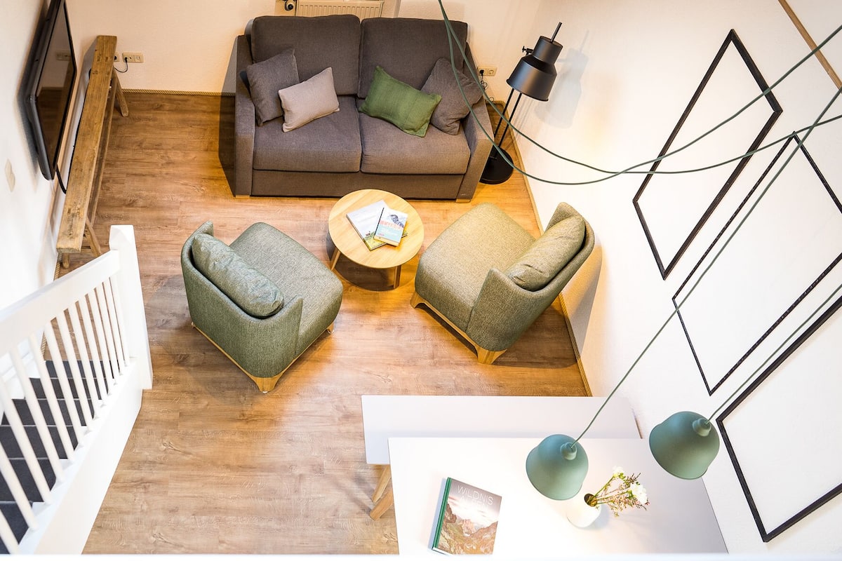 A modern living room with a grey sofa, two armchairs, and a coffee table. There is a TV mounted on the wall, a lamp, and a framed picture.