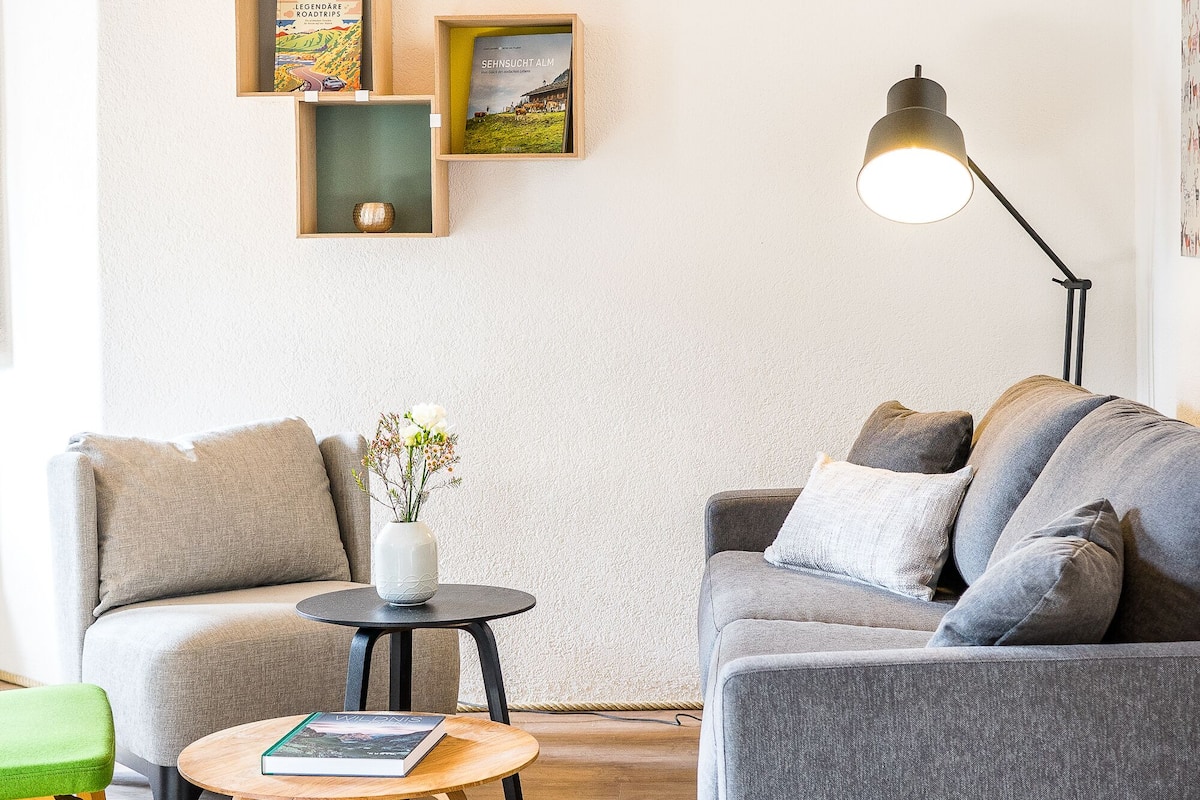 A modern living room with a grey sofa, a green chair, a small round table with a book, and a floor lamp.