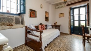 A bedroom with a stone wall, a bed with a white bedspread, a wooden chair, a table with a vase, and a window with blue shutters. - Venetiko Apartments (Naxos)