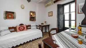 A room with a bed, a wooden table with a tablecloth, a chair, a wall clock, a small shelf with decorative items, and a window with a view. - Venetiko Apartments (Naxos)