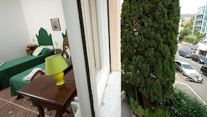 A room with a bed, a table with a lamp, and a view of a street with parked cars and a tall tree. - 2 Passi Da (Rome)