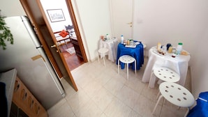A kitchen with a refrigerator, two small tables with blue and white tablecloths, and a dining area with a chair and a small table. - 2 Passi Da (Rome)