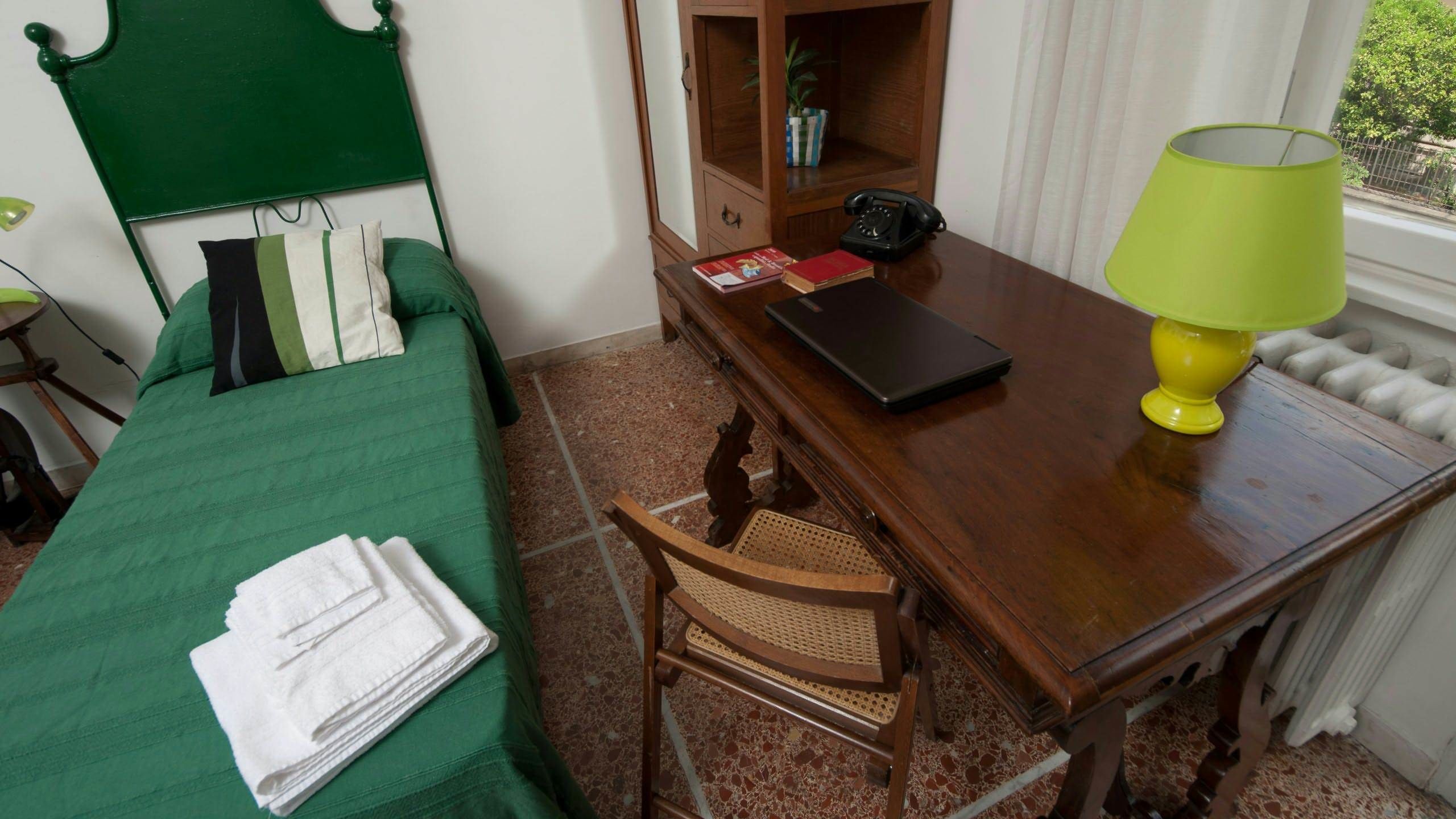 A room with a bed, a wooden desk, a chair, a lamp, and a radiator.