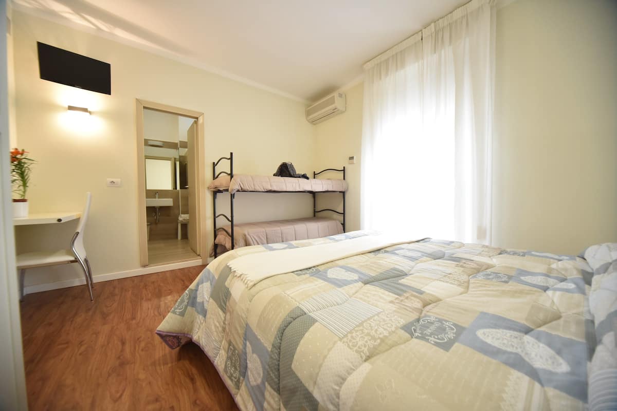 A bedroom with a bunk bed, a desk with a chair, a bedside table, and a window with curtains.