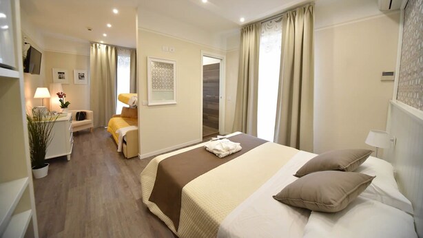 A hotel room with a bed, bedside table, lamp, and a small seating area with a chair and table. - Villa Edda (Riccione)