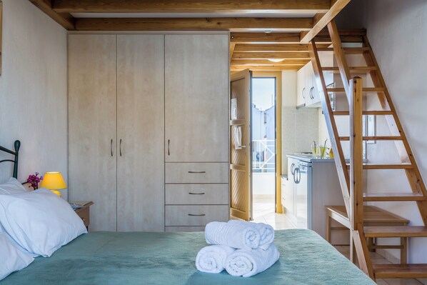 A bedroom with a bed, a wooden wardrobe, a wooden ladder, and a view into the kitchen.