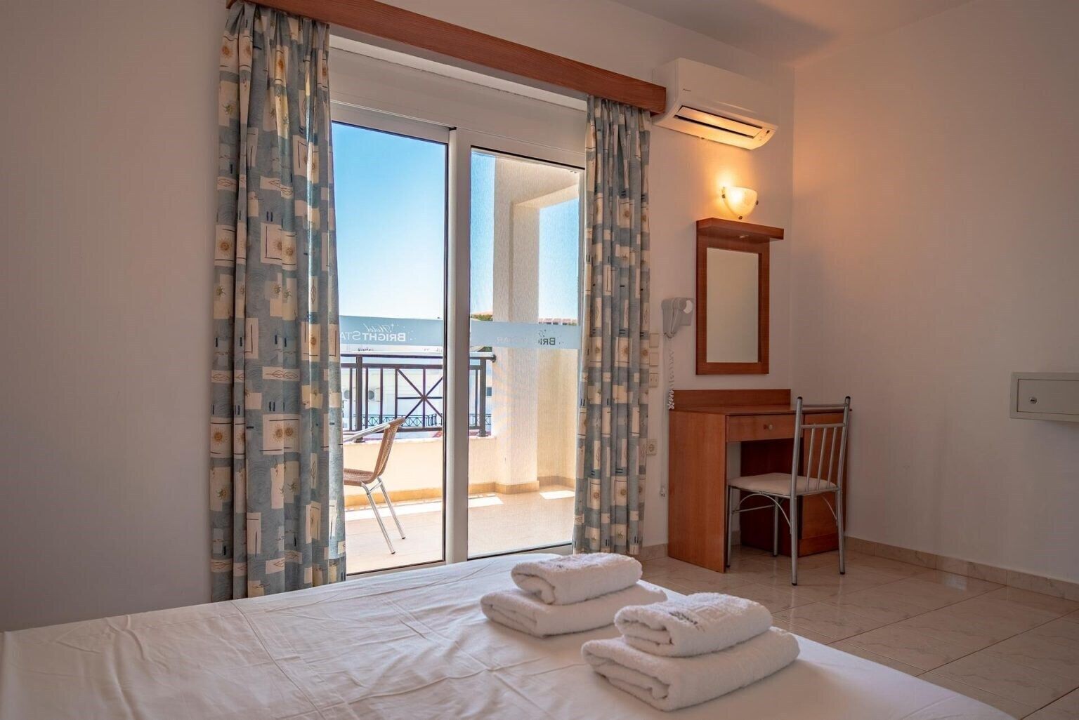 Habitación de hotel con cama, escritorio, silla y balcón con vistas.