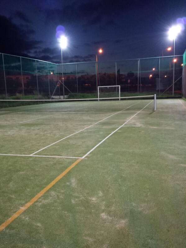 Tenniskenttä, jossa on verkko, ja jonka ympärillä on aita. Kenttää valaistaan tehokkailla valonheittimillä.