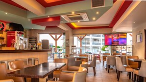 Un restaurant moderne avec des tables hautes et des chaises rembourrées, un coin bar doté d’un réfrigérateur, et de grandes fenêtres offrant une vue sur la ville.