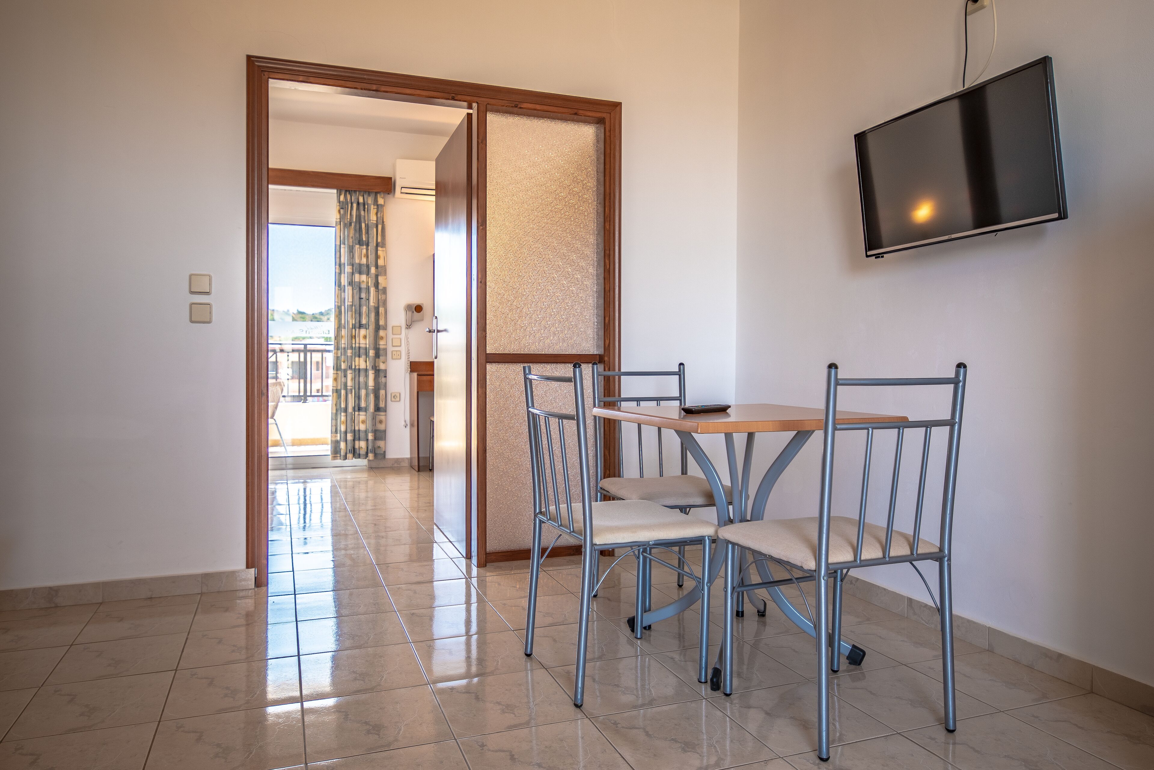 Una habitación con una mesa de comedor y sillas, un televisor montado en la pared, y vistas a un balcón.
