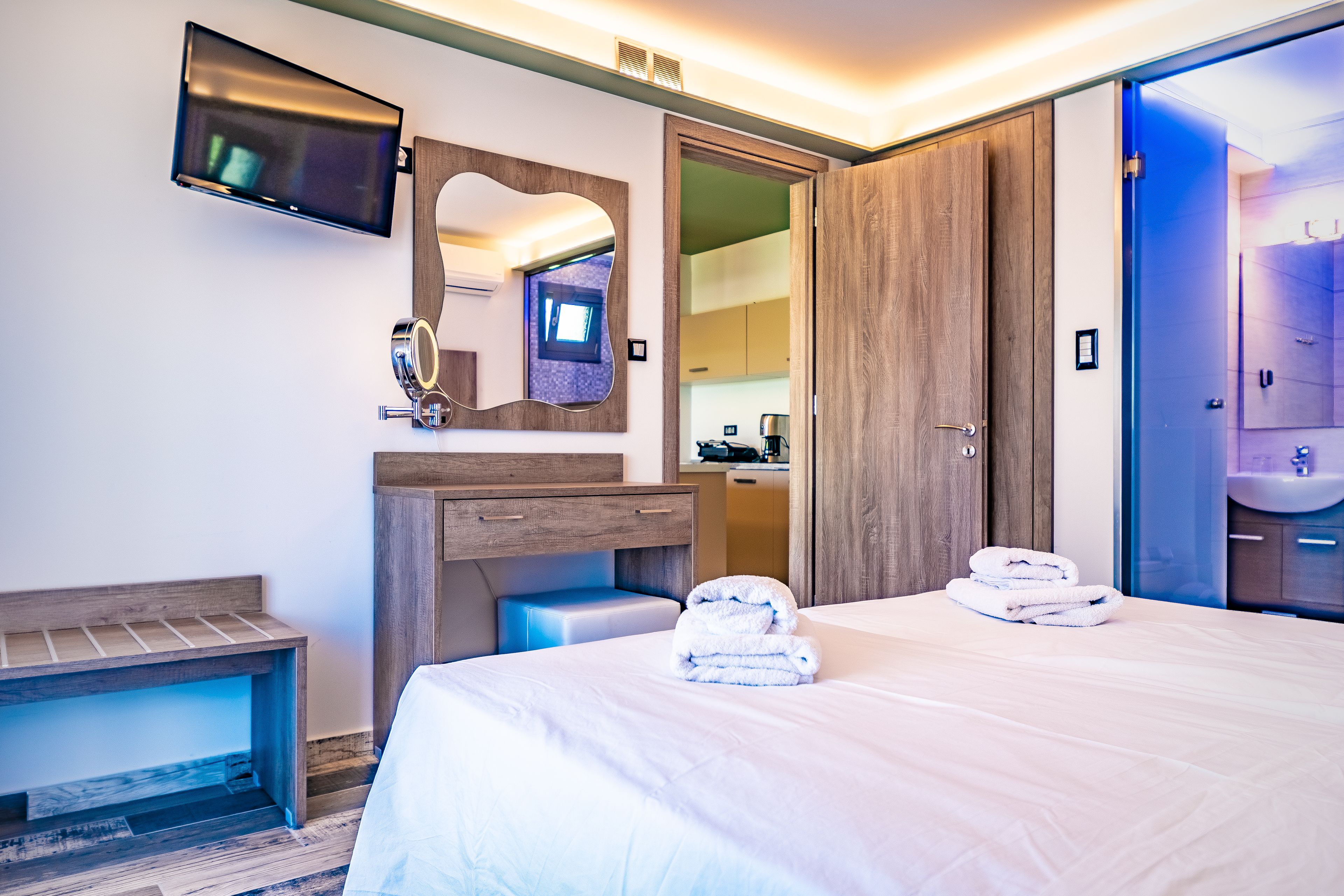 Habitación de hotel con una cama, una tocador de madera, un espejo, un televisor y un baño visible a través de una puerta abierta.