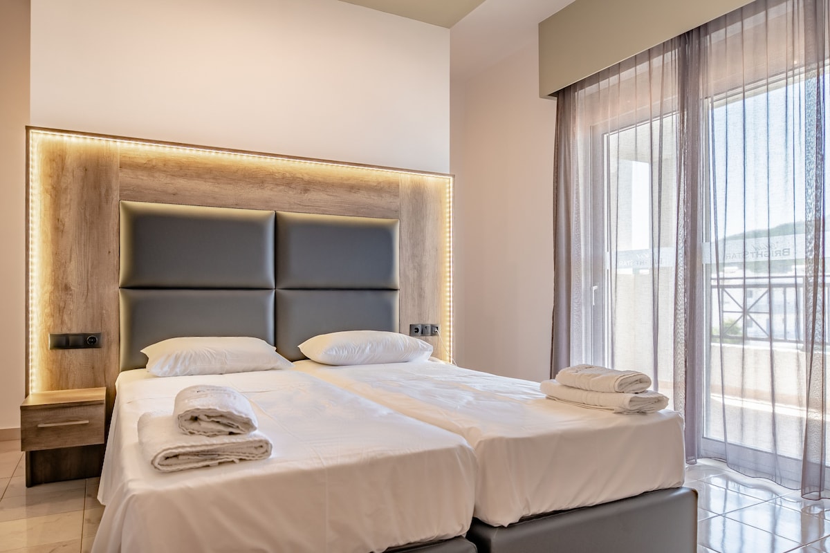 A modern hotel room with a large bed, bedside tables, and a view through sheer curtains.