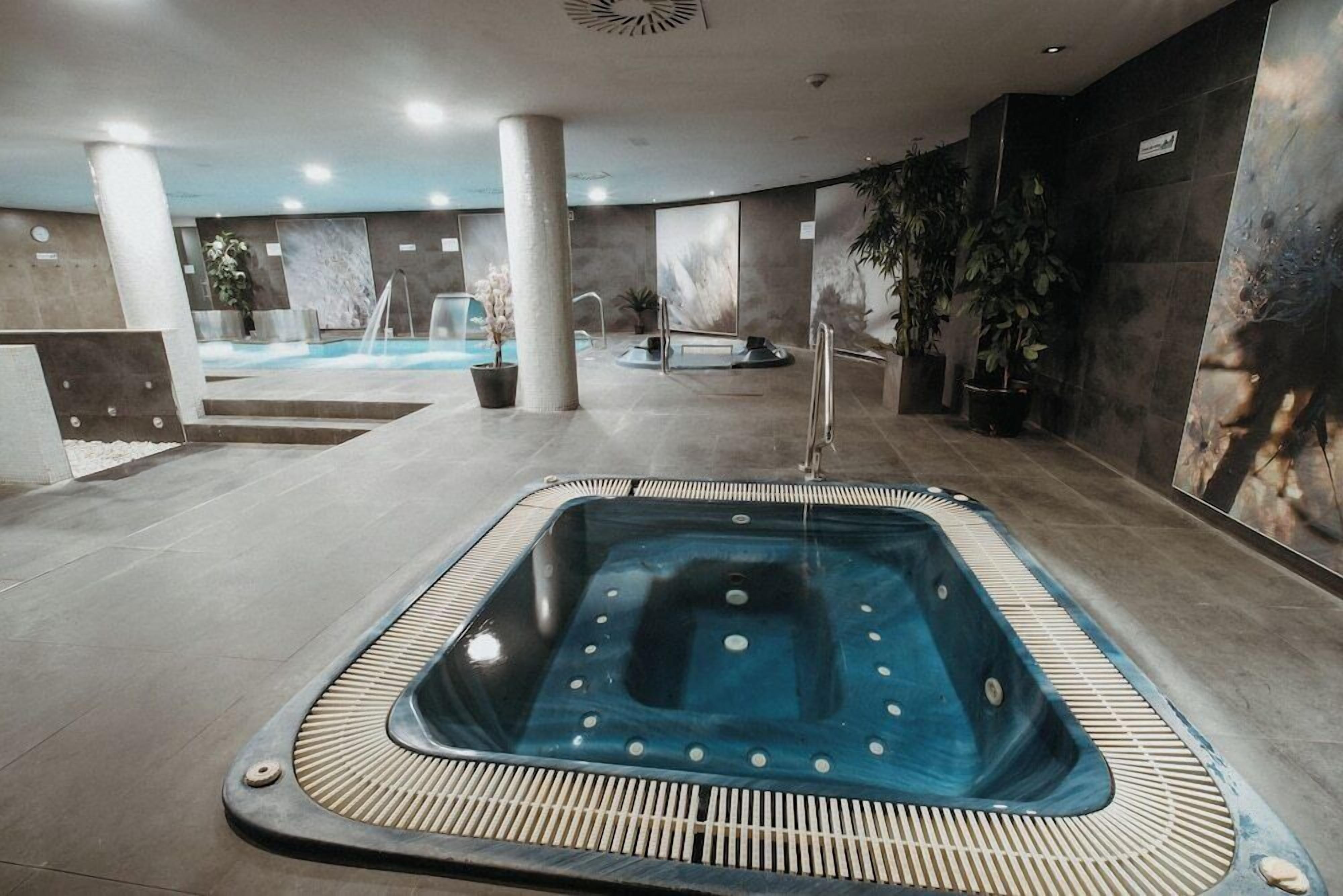 A modern indoor swimming pool with a glass enclosure, surrounded by pillars and potted plants.