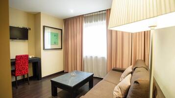 Habitación de hotel con sofá, mesa de centro, una silla roja, televisor de pantalla plana en la pared y ventana con cortinas.