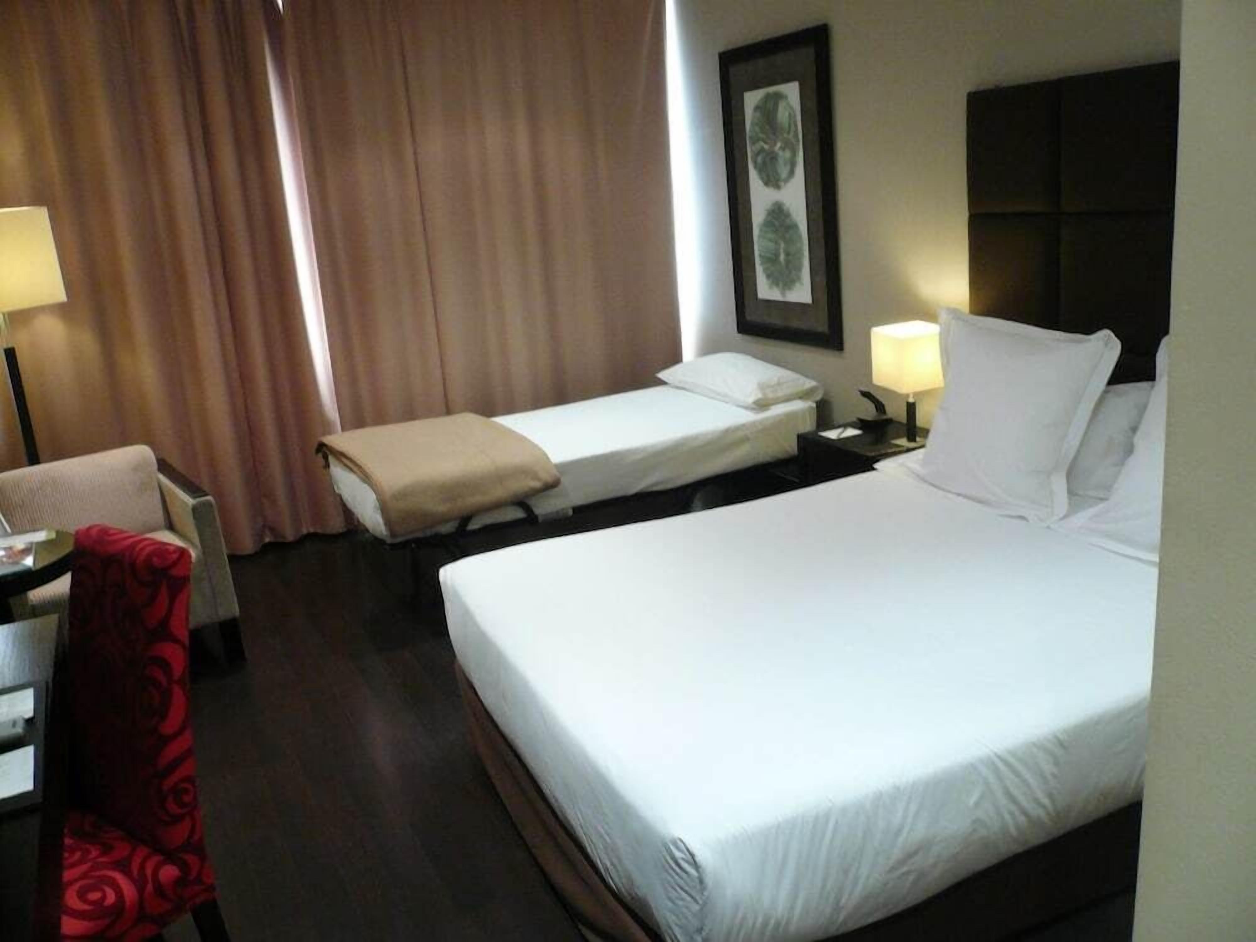 Habitación de hotel con una cama grande, una cama más pequeña, un escritorio con silla, una lámpara y un cuadro en la pared.