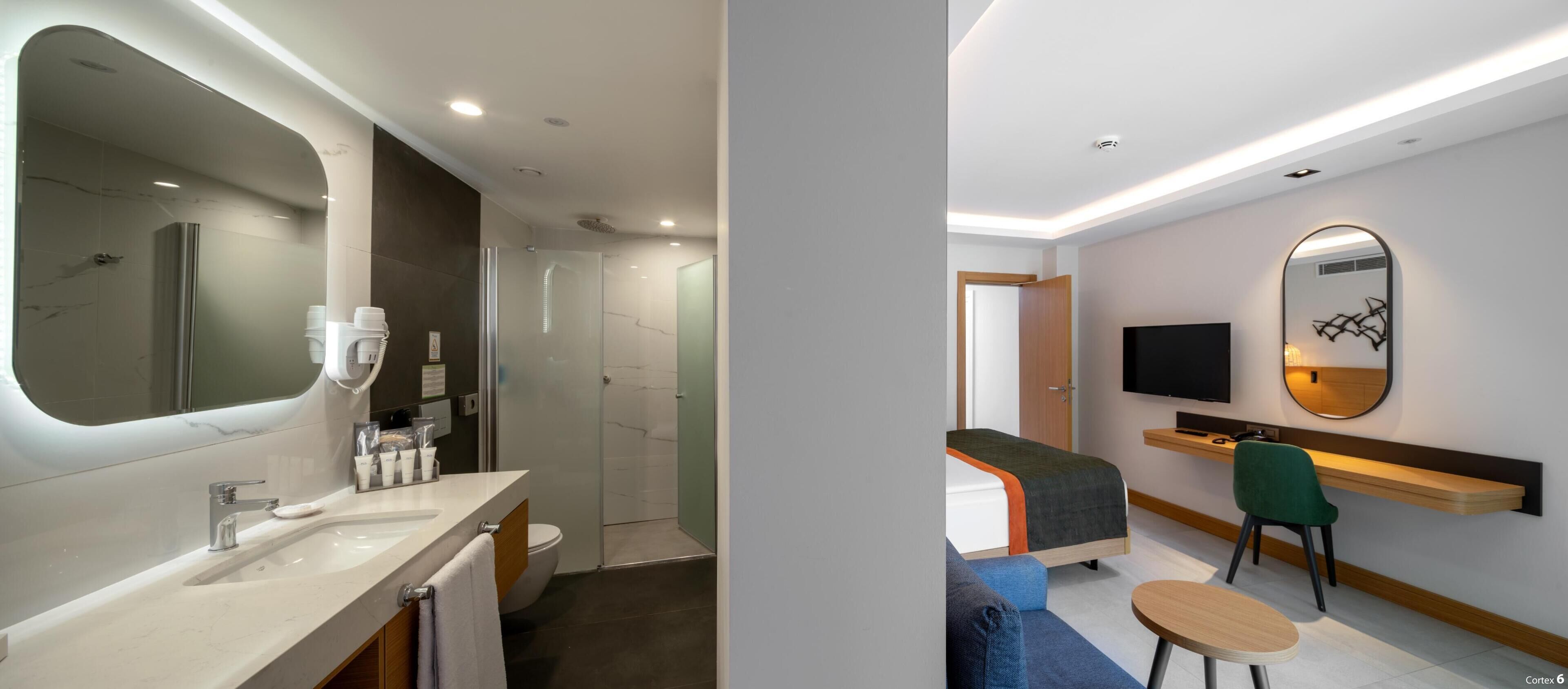 A modern bathroom with a large mirror, sink, and shower. The room features a bed, a TV, and a desk with a chair.