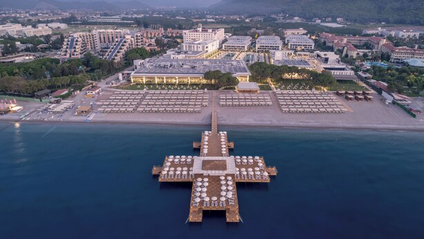 An aerial view of a coastal hotel with a long pier extending into the sea, featuring multiple dining areas with tables and chairs.