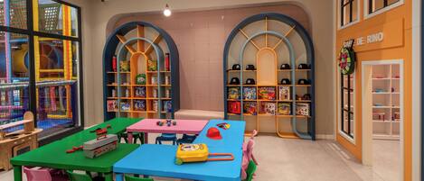 A children's play area with colorful tables and chairs, shelves with toys, and a large window with a view of an outdoor play structure.