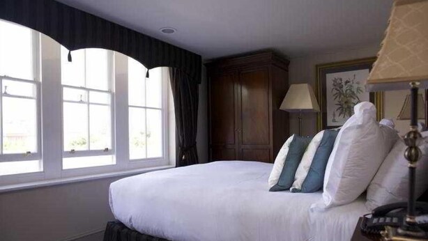 A hotel room with a large bed, a window with curtains, a wooden cabinet, and a lamp. - The Max Brighton (Brighton)