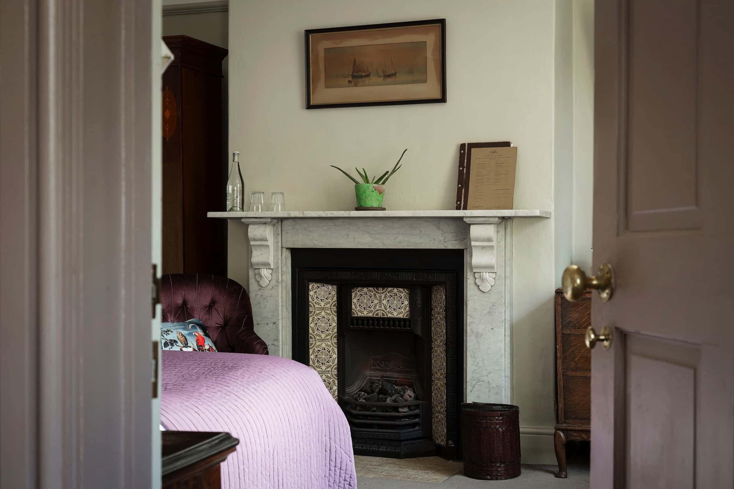 A room with a fireplace, a bed with a purple blanket, a green plant on the mantel, and a framed picture on the wall.