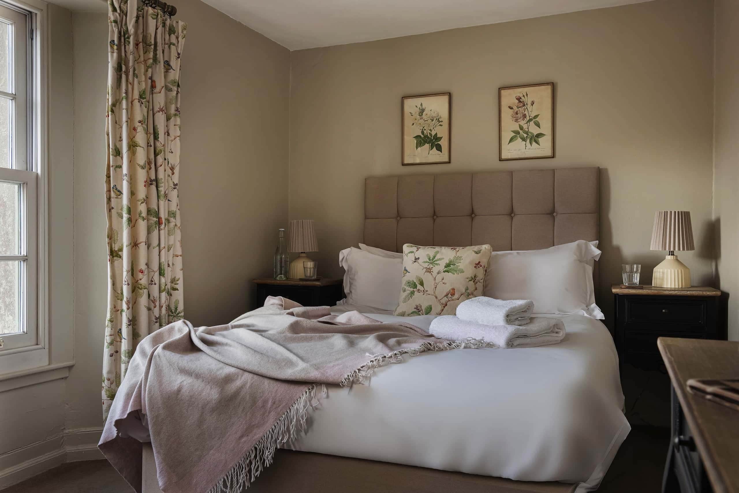 Una cama bien hecha con un cabecero color beige, una mesita de noche con una lámpara y una ventana con cortinas.