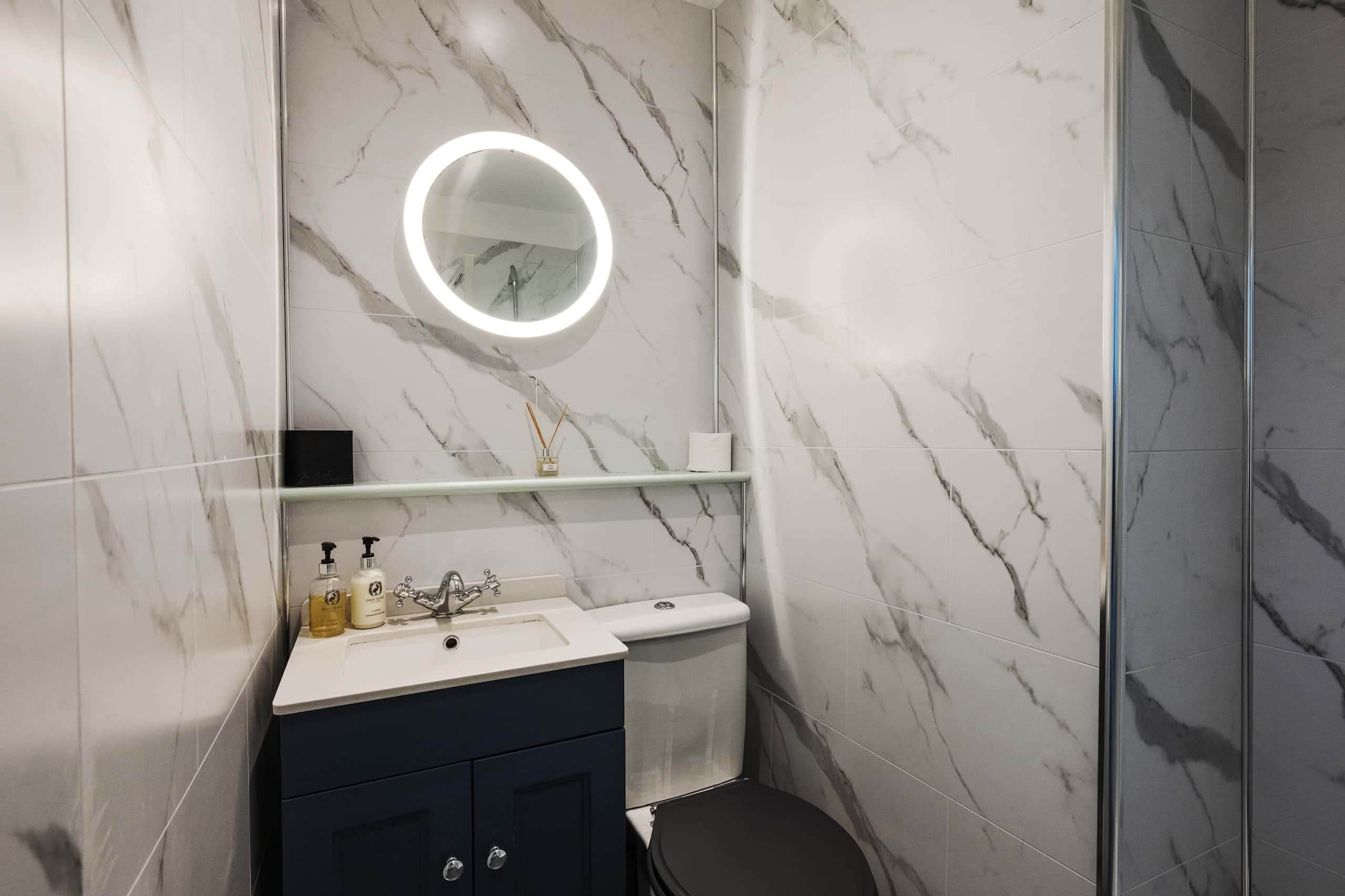 Un baño con pared de mármol, un espejo redondo con luz, un lavamanos y un inodoro.