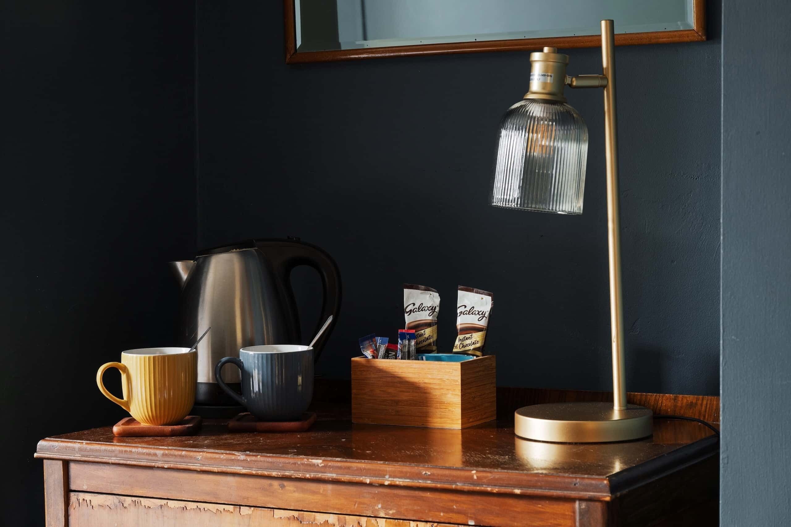 A wooden table with a coffee maker, two cups, a box of instant coffee, and a floor lamp.