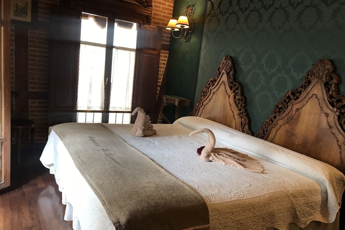 Quarto com cama grande, duas almofadas, cabeceira de madeira, janela com cortinas e uma luminária.